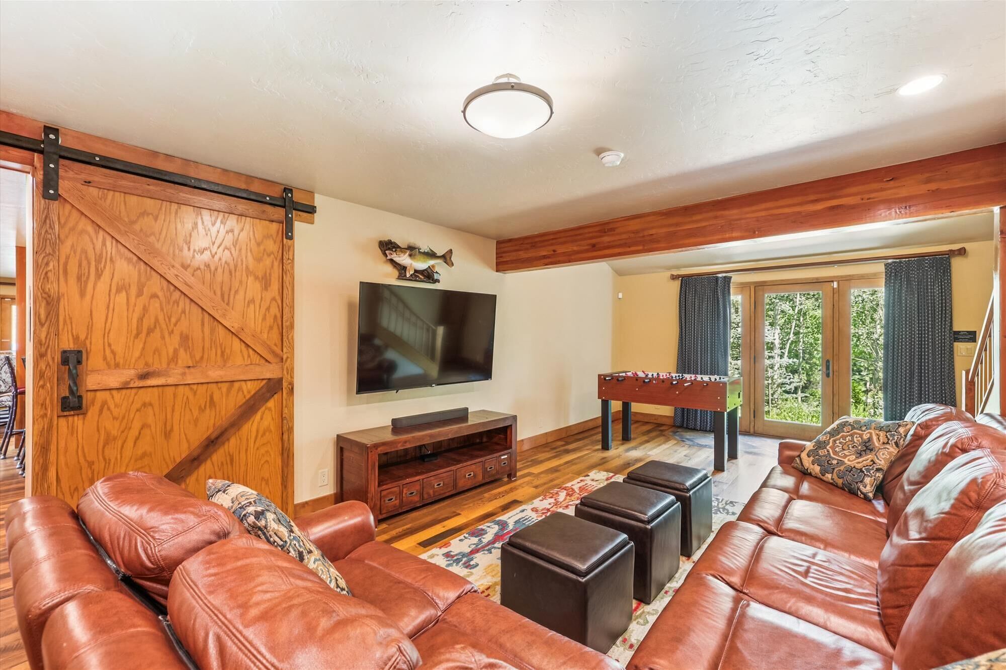 A spacious living room with a large leather sofa, a flat-screen TV mounted on the wall, a wooden coffee table, and a foosball table.
