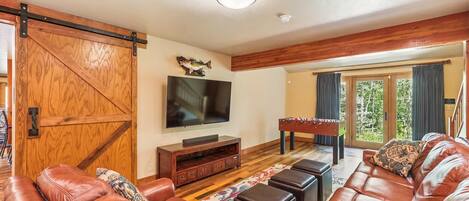A spacious living room with a large leather sofa, a flat-screen TV mounted on the wall, a wooden coffee table, and a foosball table.