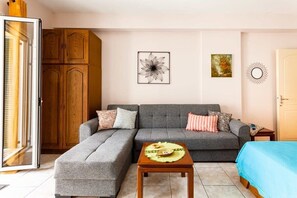 A living room with a grey sofa, a wooden cabinet, a small table with a placemat, and a blue bed. - Kalamata City Nest - Central & Cozy Hideout (Kalamata)