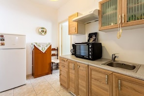 A compact kitchen with a white refrigerator, a microwave, a sink, wooden cabinets, and a small dining area with a chair and table. - Kalamata City Nest - Central & Cozy Hideout (Kalamata)