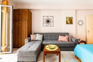 A living room with a grey sofa, a wooden cabinet, a small table with a decorative item, and a bed with a blue cover.