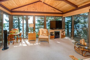 A cozy room with a TV, wicker furniture, a fireplace, and large windows overlooking a forested area.