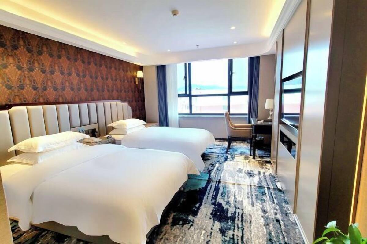 A hotel room with two beds, a desk, a chair, a large window, and a patterned carpet.