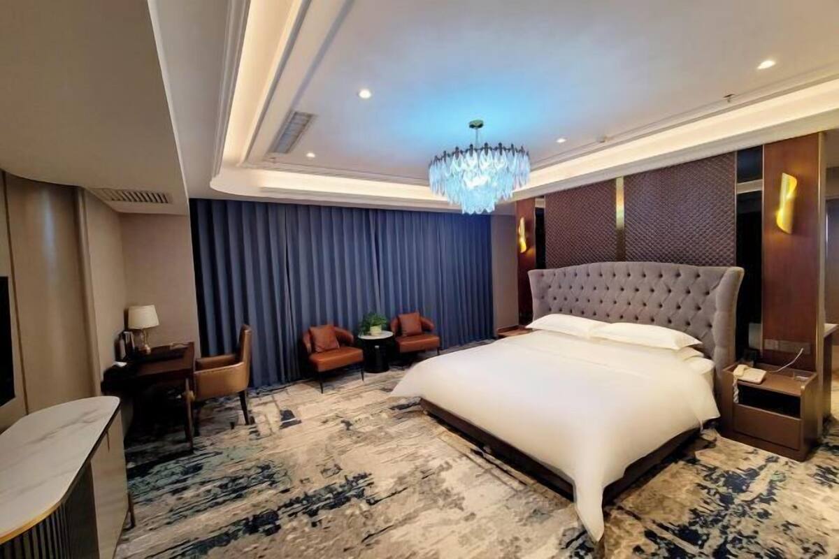 A hotel room with a large bed, a chandelier, a desk with a lamp, two chairs, and a small table with a plant.