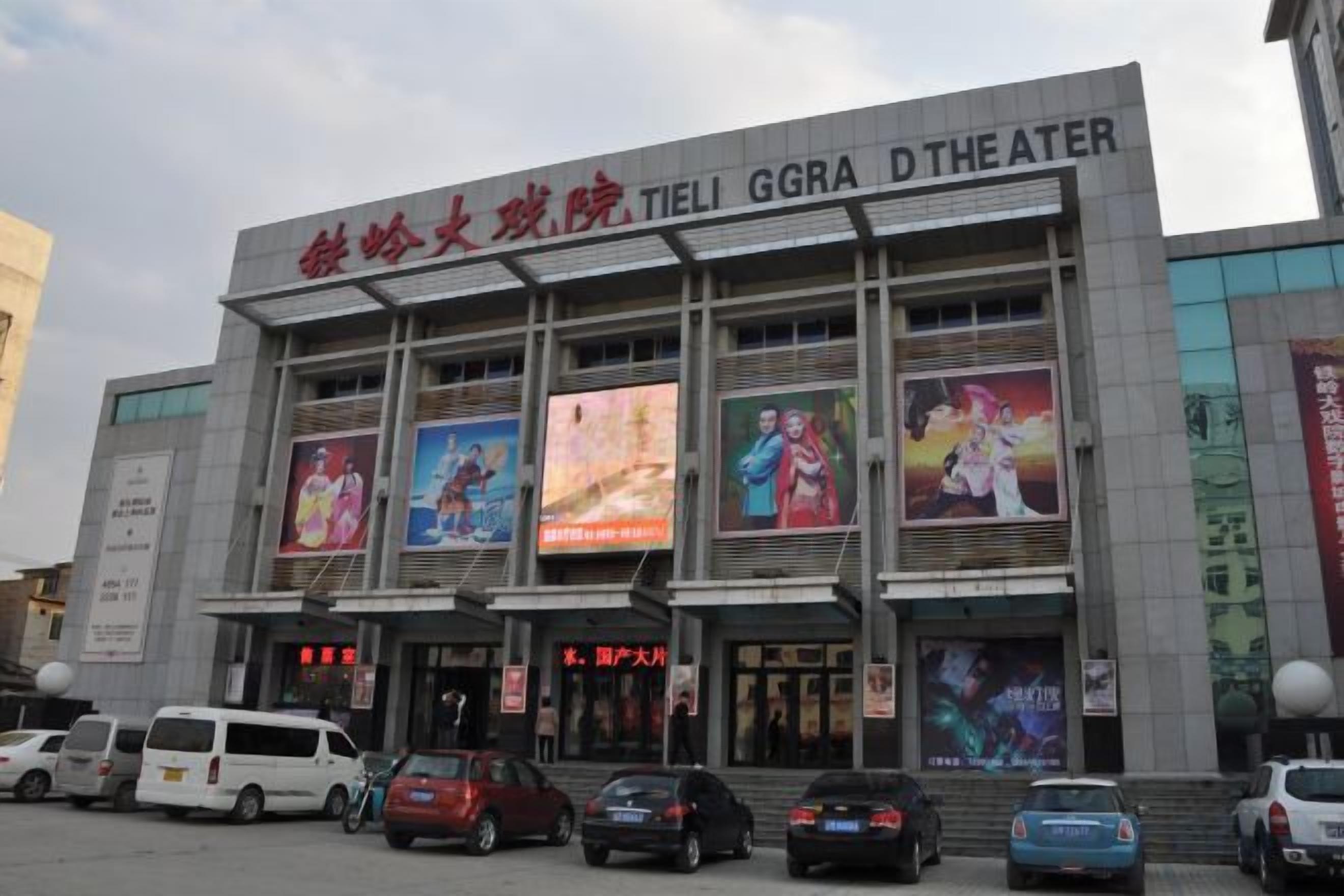 A theater building with multiple large posters on the facade, a sign indicating it is the Tieli GGRA Theater, and several parked cars in front.