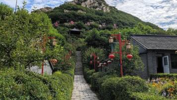 A stone pathway leading to a traditional building with red lanterns, surrounded by lush greenery and a mountain backdrop.