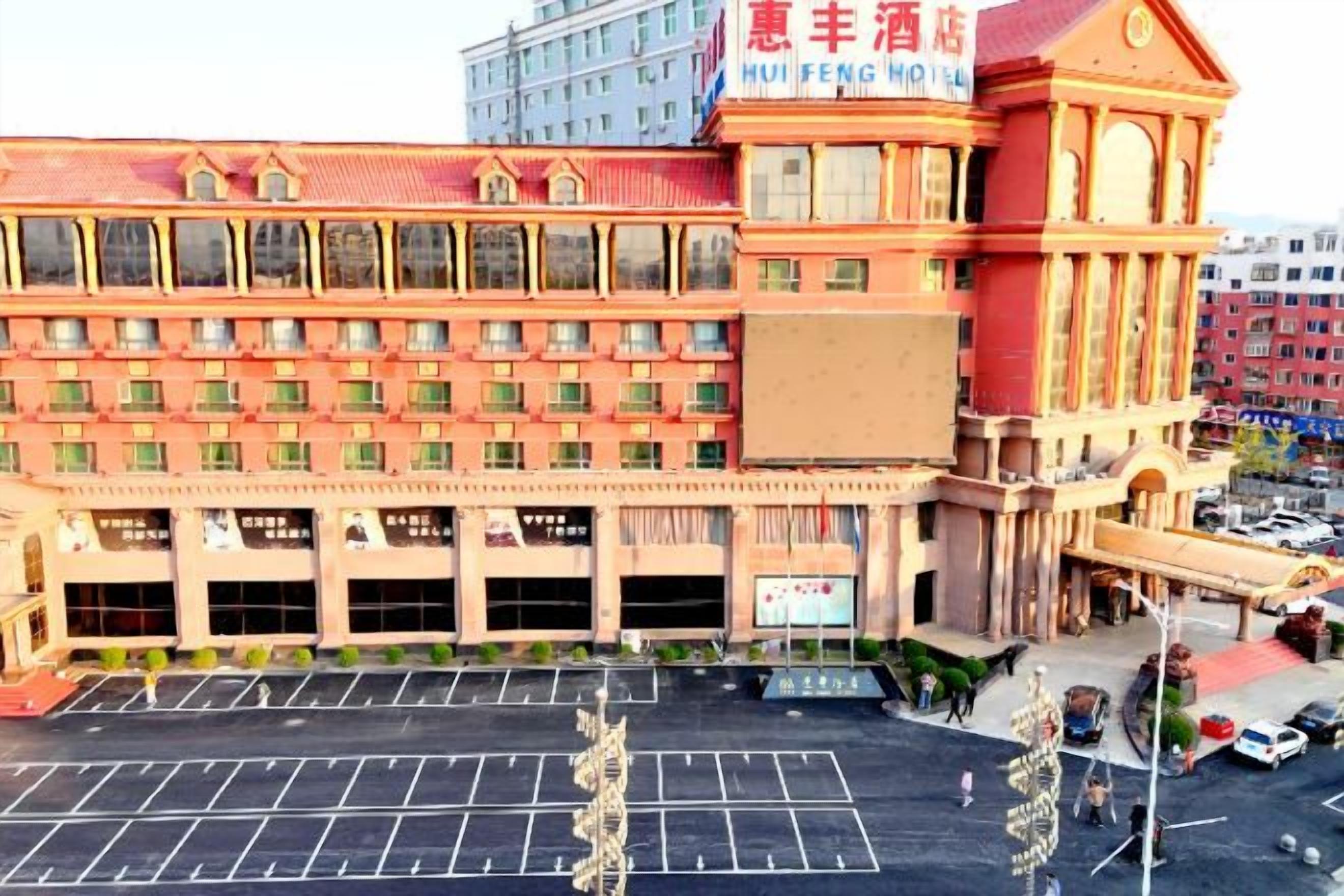Un hotel a più piani con una facciata imponente in rosso e beige, diversi posti auto e un grande cartello con la scritta 'HUI FENG HOTEL'.