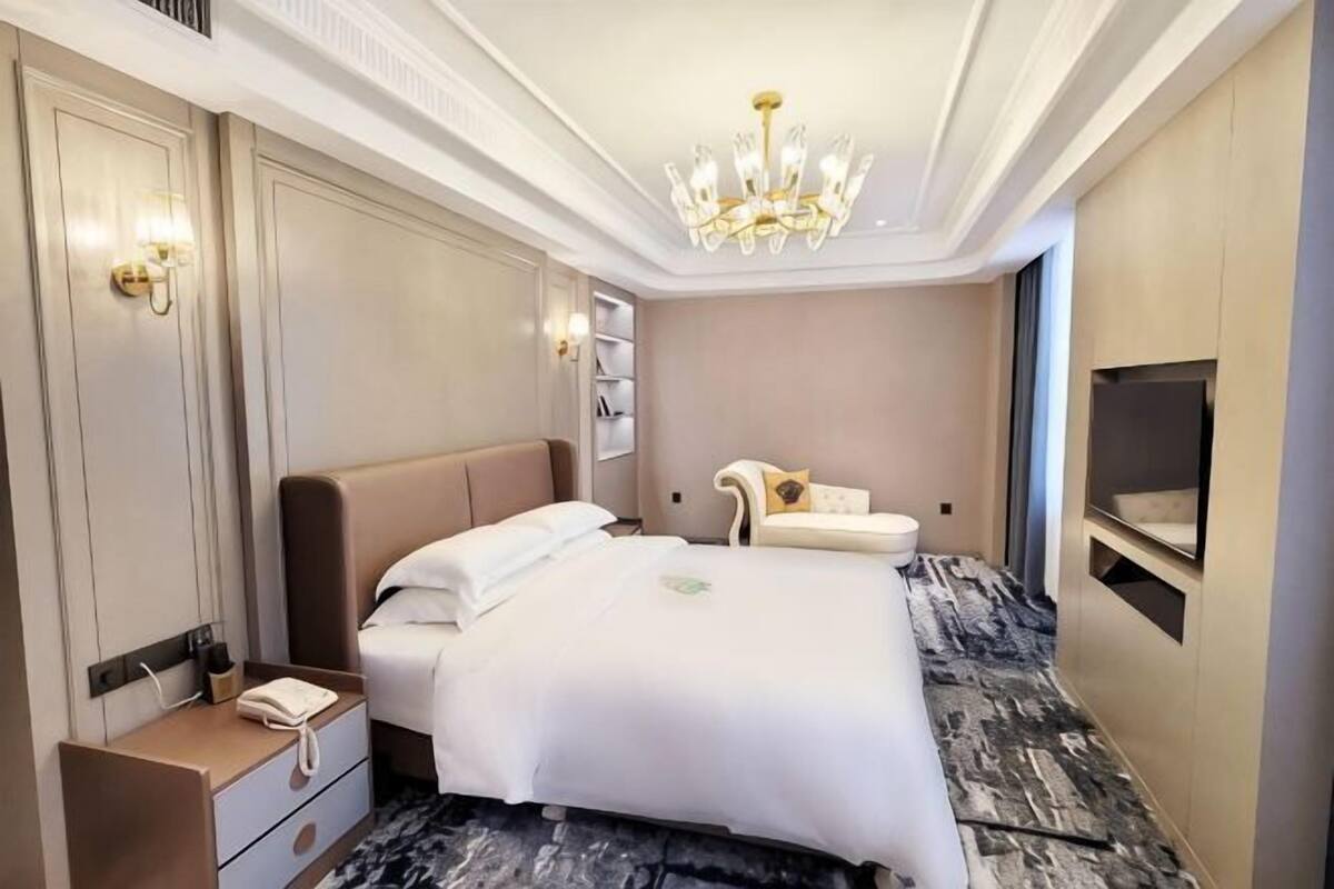 A hotel room with a large bed, a bedside table, a chair, a TV, and a chandelier.