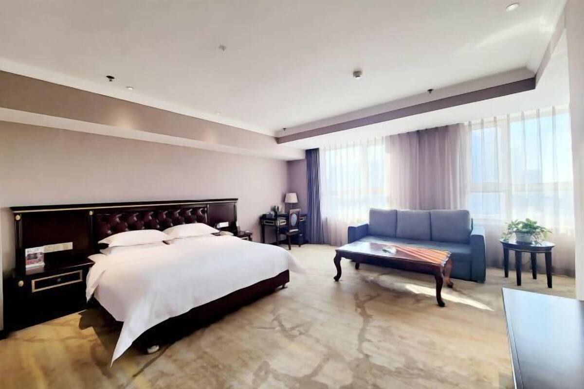 A modern hotel room with a large bed, a sofa, a coffee table, and a desk.
