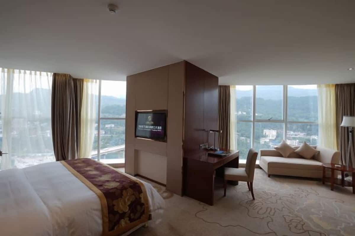 Een hotelkamer met een bed, een bureau, een bank, een televisie en grote ramen met uitzicht over de stad.