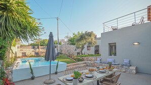 A poolside dining area with a table set for two, surrounded by wicker chairs, a stone wall, and a view of a swimming pool. - Villa Garifalia with Sauna & Hydromassage (Apokoronas)