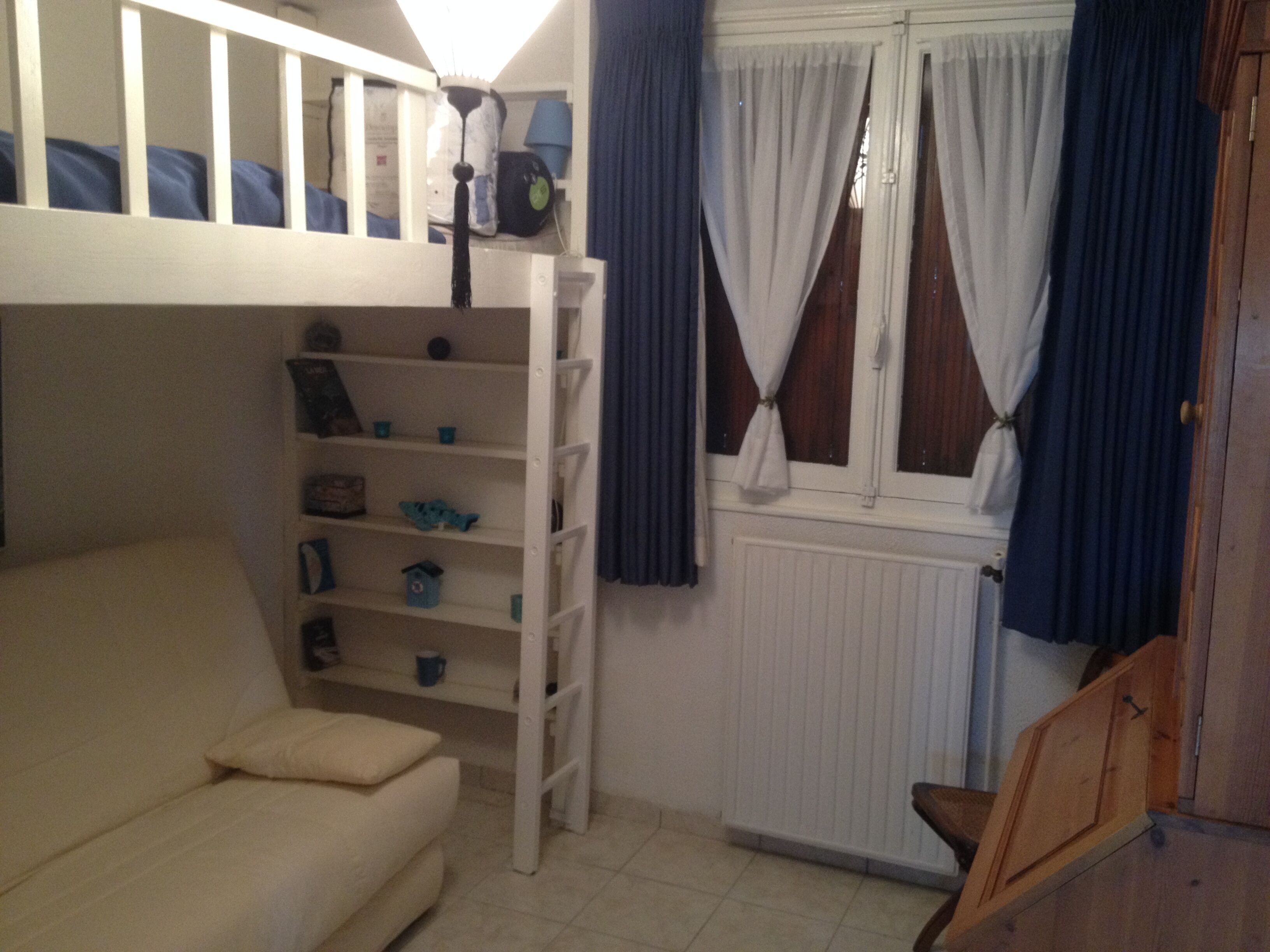 Een kamer met een stapelbed, een slaapbank, een radiator en een raam met gordijnen.