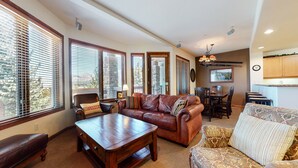 A spacious living room with a leather sofa, a wooden coffee table, and a patterned armchair. There is a dining area with a table and chairs, and a kitchen area with wooden cabinets. - Eagle Run #114 2 Bedroom Condo by RedAwning (Mammoth Lakes)