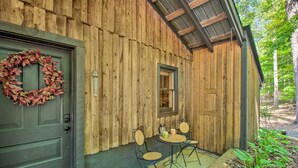 Uma cabana rústica de madeira com porta verde escura, uma grinalda, uma pequena janela e uma mesa com cadeiras na parte externa.