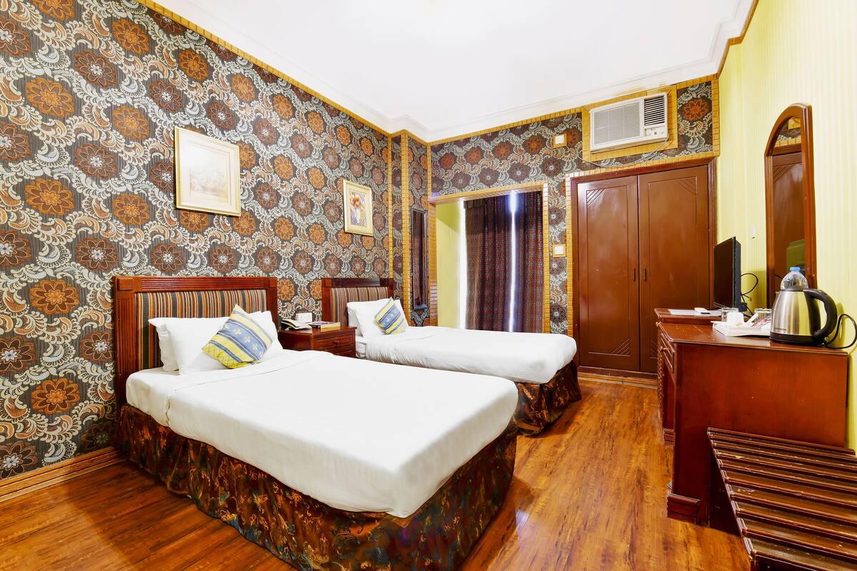 Une chambre d’hôtel avec deux lits, un plancher en bois, un papier peint à motifs, un bureau avec une bouilloire et une armoire.