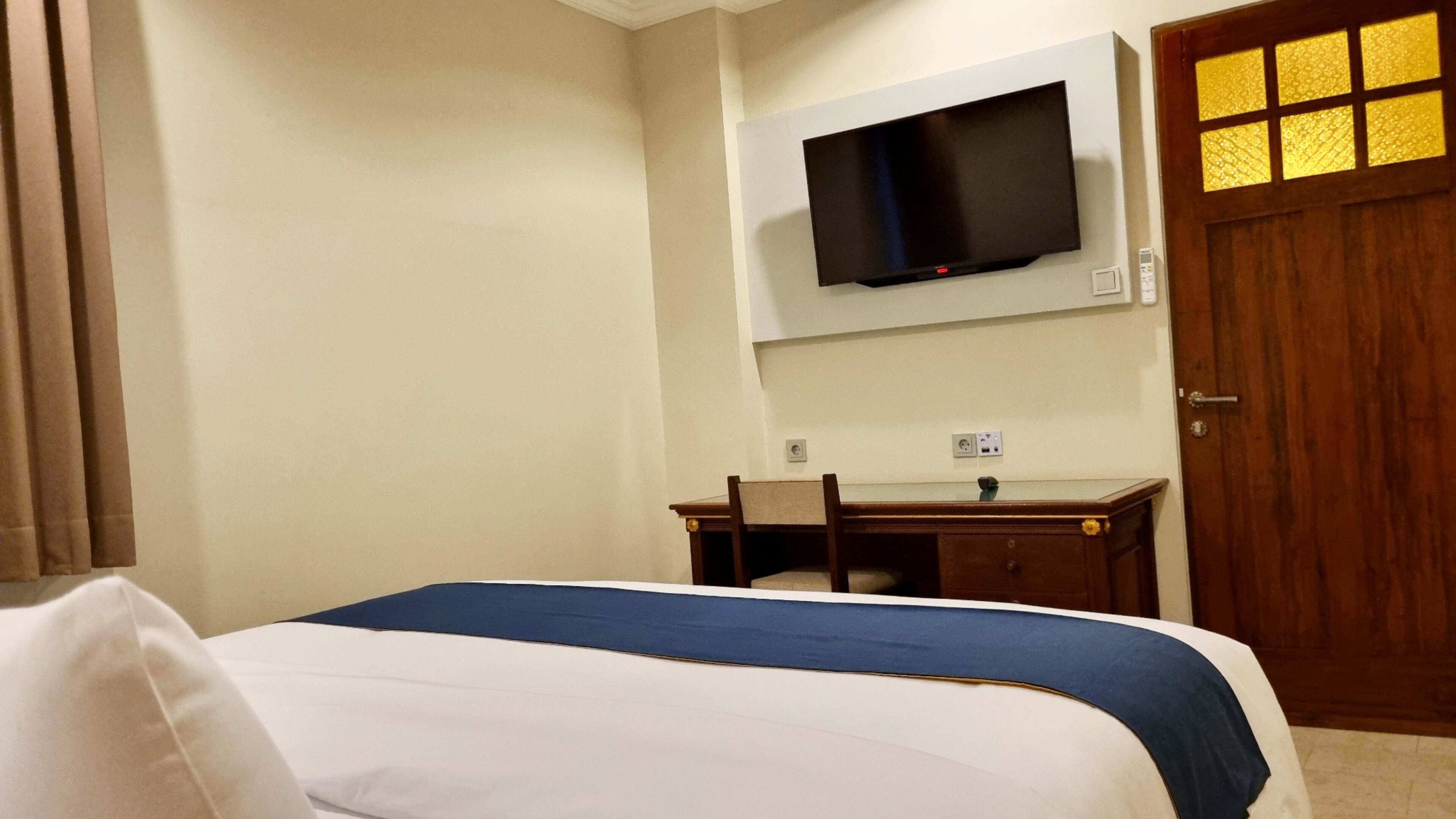 Habitación de hotel con cama, escritorio, silla, televisor montado en la pared y puerta de madera con ventanal.