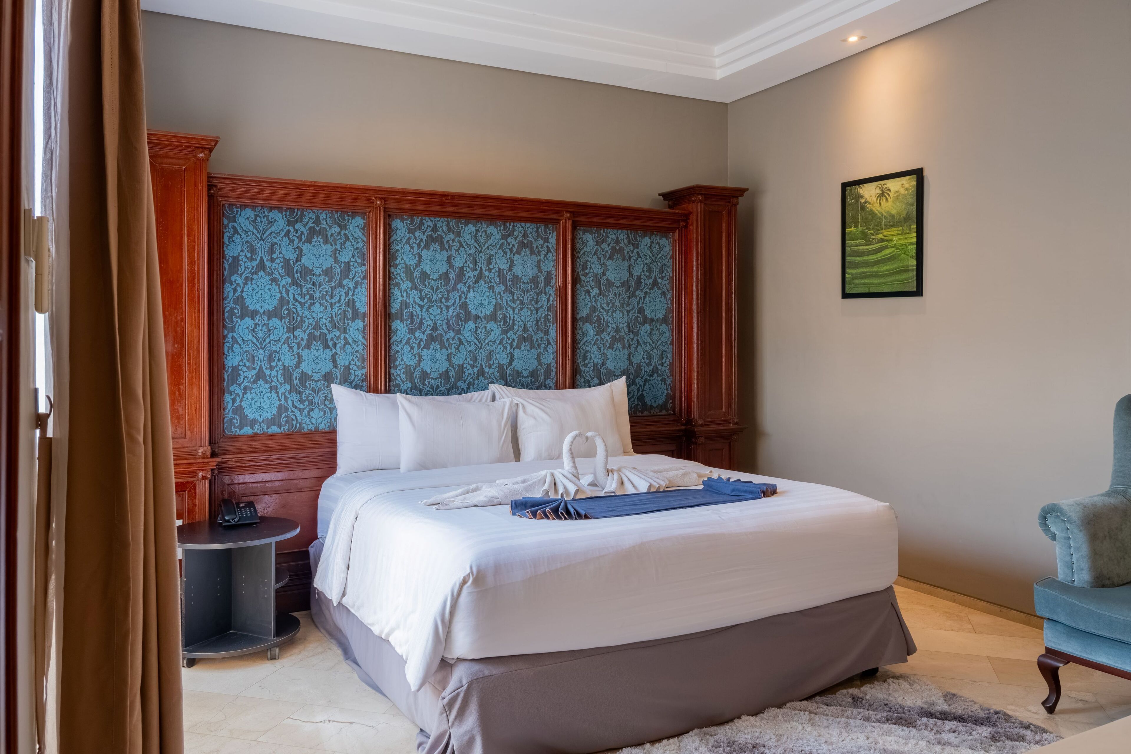 A neatly made bed with white linens and a blue towel, a wooden headboard with blue damask panels, a small bedside table with a phone, and a framed landscape painting on the wall.