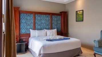 A neatly made bed with white linens and a blue towel, a wooden headboard with blue damask panels, a small bedside table with a phone, and a framed landscape painting on the wall.
