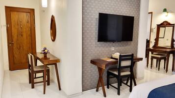 Een hotelkamer met een houten bureau, twee stoelen, een aan de muur bevestigde televisie, een spiegel en een houten deur.