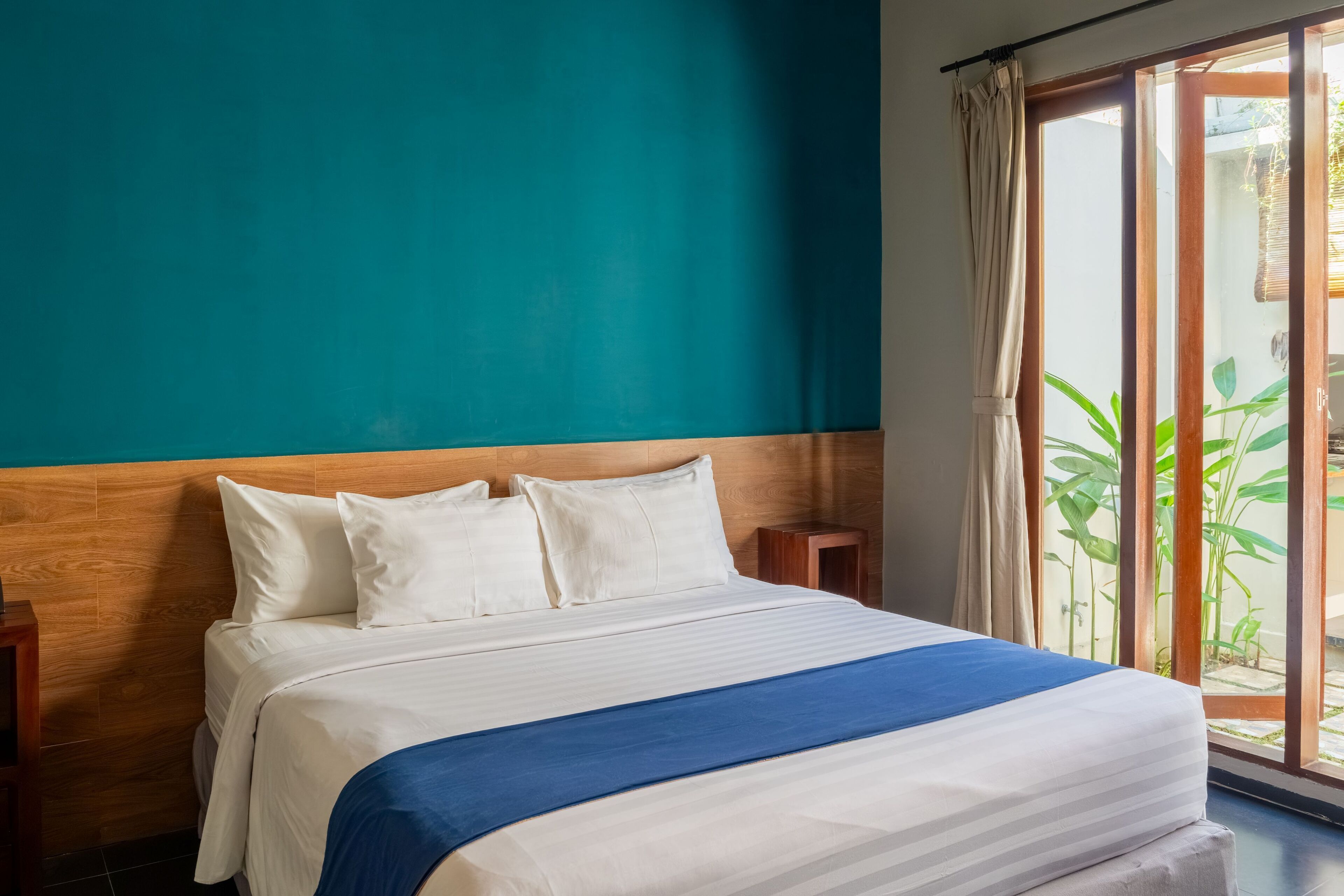 A neatly made bed with white and blue bedding, a wooden headboard, and a view of greenery through a sliding glass door.