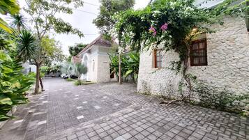 A paved pathway leading to a white building with a stone facade, surrounded by lush greenery and a tiled roof.