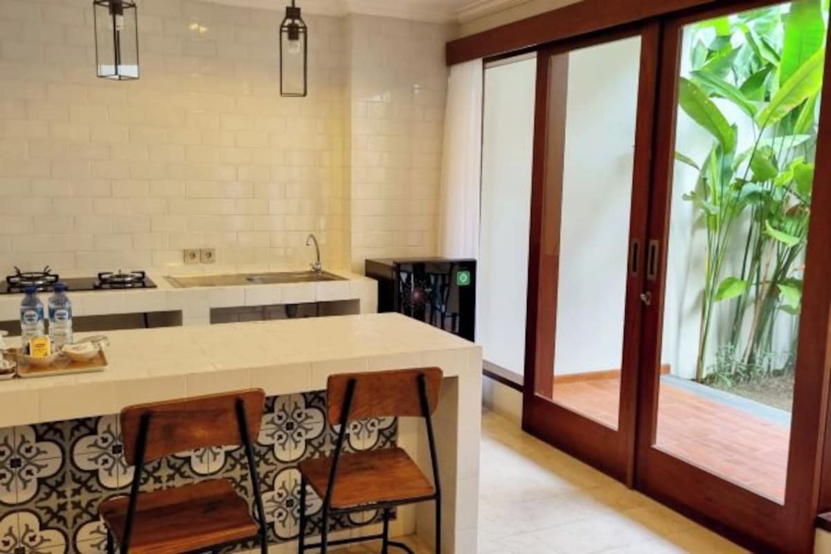A kitchen with a white countertop, two hanging lights, a sink, and a view of greenery through a sliding door.