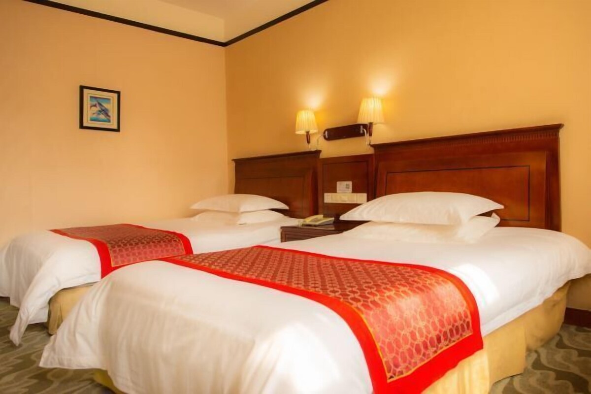 A hotel room with two beds, each with white linens and a red patterned bedspread. There are two bedside lamps and a framed picture on the wall.
