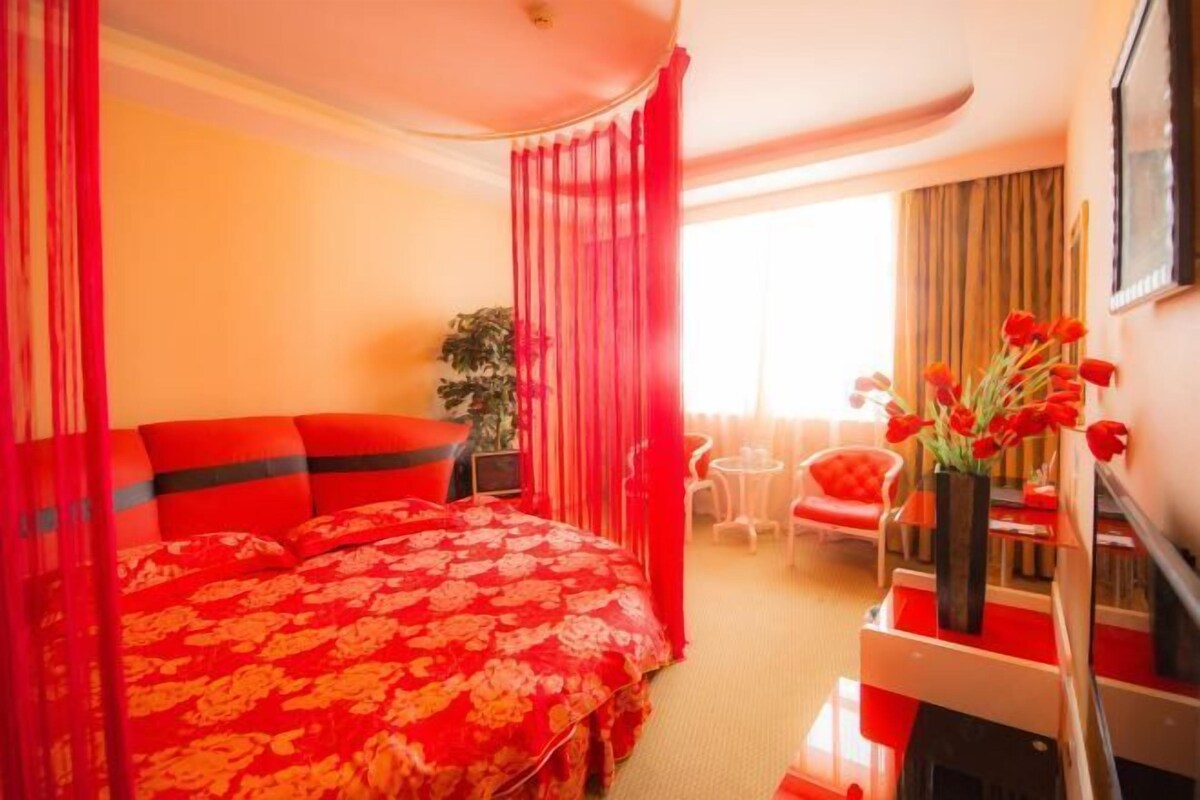 A hotel room with a large bed, red curtains, a small table with a chair, and a vase of red flowers.