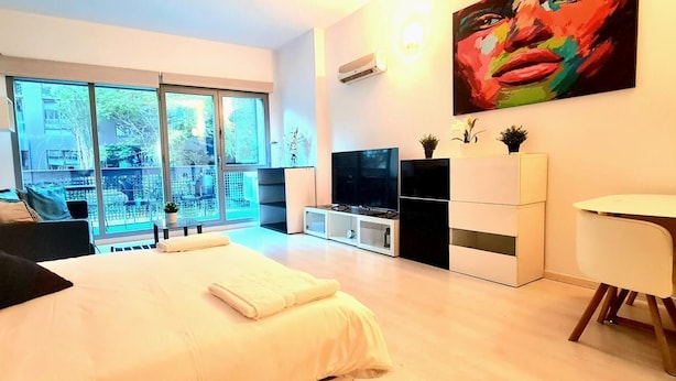 A modern hotel room with a large bed, a TV, a dining area, and a balcony with a view.