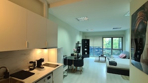 A modern kitchen with a dining area, a living room with a sofa and a large window, and a balcony with a view.