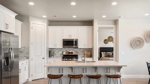A modern kitchen with white cabinets, a stainless steel refrigerator, a microwave, and a gas stove. There are three wooden stools at the island. - A Family Affair, Sleeps 20 in two side-by-side units, 10 bedrooms! (Washington)
