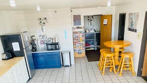 A clean, tiled floor with a kitchen area featuring blue cabinets, a refrigerator, and a coffee machine. There is a wooden table with four stools in the center. - Home Place Inn (Nicholasville)
