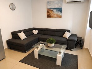 Ein modernes Wohnzimmer mit einer Eckcouch, einem Glastisch, einem kleinen Topfpflanzen und einem Gemälde von einer Strandlandschaft.