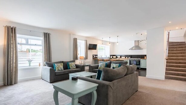 A modern living room with a grey sofa, a small table, and a kitchen in the background. - 5 Bedrooms, Sleeps 10-12 w/ Wifi, Parking & Garden (Leven)