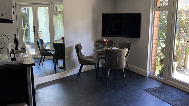 A modern living room with a dining area, a flat-screen TV mounted on the wall, and a large sliding glass door leading to a garden. - Thistle Lodge - Quiet Garden Lodge with off road parking (Hemel Hempstead)