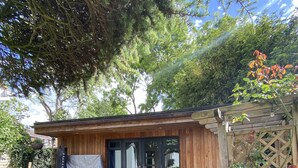 A wooden building with a glass door, surrounded by potted plants and a stone patio. - Thistle Lodge - Quiet Garden Lodge with off road parking (Hemel Hempstead)