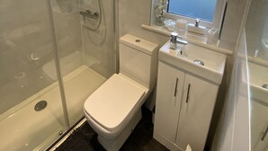 A bathroom with a glass-enclosed shower, a toilet, a sink with a window above, and a small shelf with toiletries. - Thistle Lodge - Quiet Garden Lodge with off road parking (Hemel Hempstead)