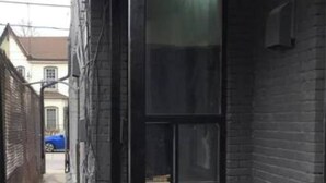 Un callejón estrecho con un edificio negro a la derecha, que tiene un gran ventanal y una puerta pequeña.