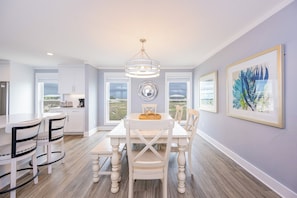 A dining area with a white table, chairs, and a chandelier. There are windows with a view of the outdoors.