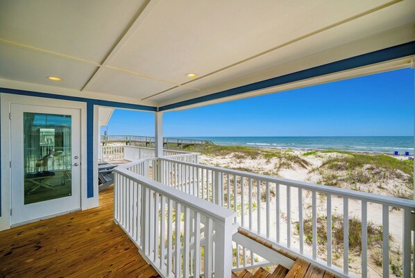A wooden deck with a white railing, a glass door, and a view of the beach and ocean.