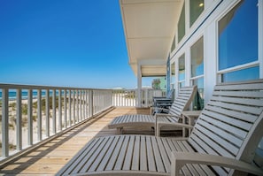 En træterrasse med hvide gelændere, udendørs møbler og udsigt til stranden.