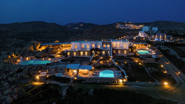 A luxurious resort with multiple swimming pools, a well-lit main building, and a scenic mountainous backdrop. - Villa Super Three (Mykonos)