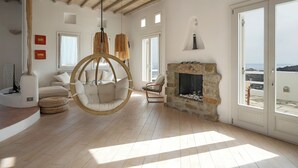 A spacious living room with a stone fireplace, a hanging chair, and a view of the sea through large windows. - Villa Super Three (Mykonos)