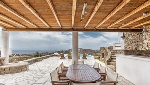 A covered outdoor seating area with a wooden table and chairs, stone flooring, and a view of the sea. - Villa Super Three (Mykonos)