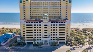 Un hôtel à plusieurs étages, doté d’un dôme central, situé près d’une plage bordée de palmiers et offrant une vue dégagée sur l’océan.