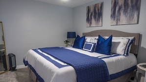 A neatly made bed with blue and white bedding, a bedside table with a lamp, and abstract wall art. - Roanoke’s modern Lower Level Apt. (Basement). (Roanoke)