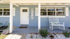 A house with a blue exterior, a white door, and a 'WELCOME' sign. There is a white rocking chair outside.