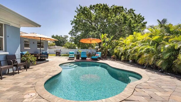 A swimming pool with a curved edge, surrounded by a paved patio with lounge chairs and umbrellas.