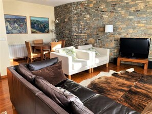 A living room with a stone wall, a leather sofa, a white sofa, a TV on a stand, and a dining area with a table and chairs. - Townhouse 4 rooms Pool, Fireplace, Wi-Fi & Pet-Friendly (Llívia)
