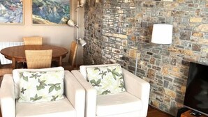 A living room with two white armchairs with leaf-patterned cushions, a wooden dining table with wicker chairs, a stone wall, and a floor lamp. - Townhouse 4 rooms Pool, Fireplace, Wi-Fi & Pet-Friendly (Llívia)